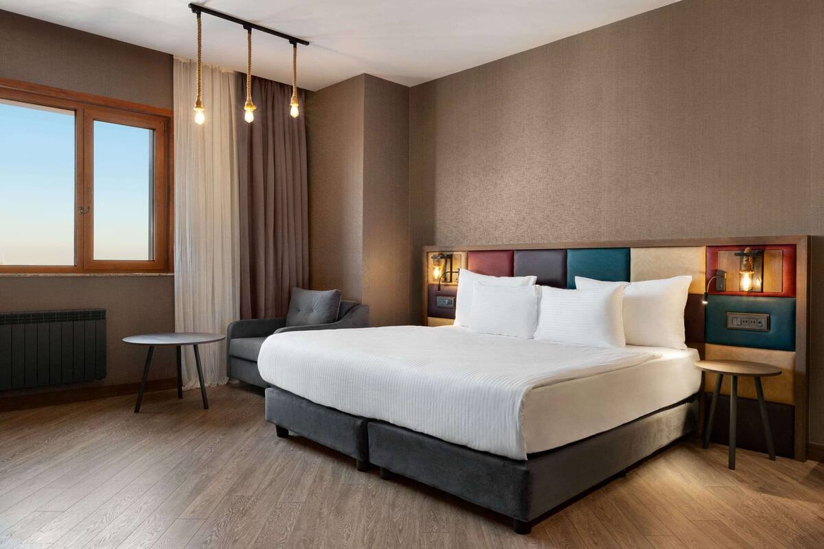 A modern hotel room with a large bed, a small table, a sofa, and a view of the outdoors.