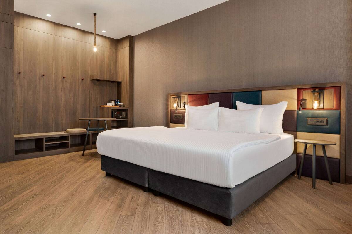 A modern hotel room with a large bed, wooden panel walls, a small table, and a bench.