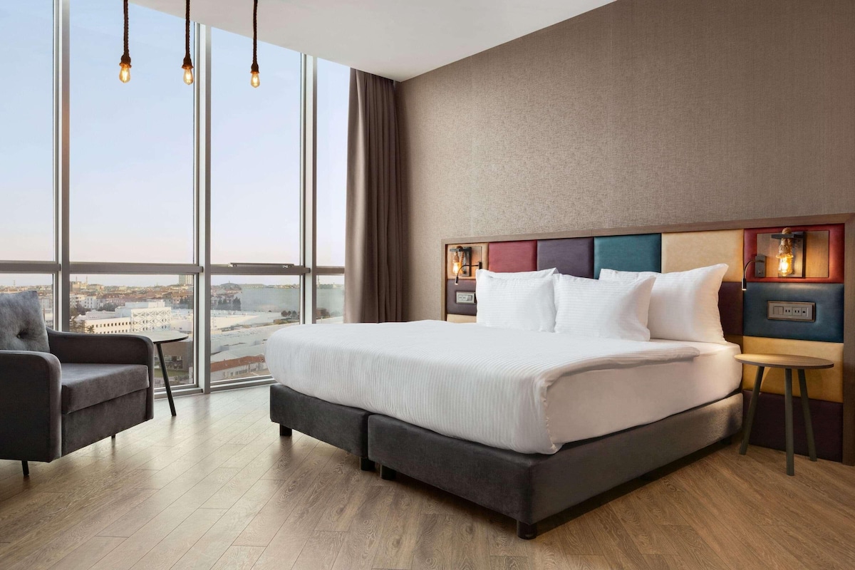 A modern hotel room with a large bed, a small table, and a chair. The room has large windows offering a city view.