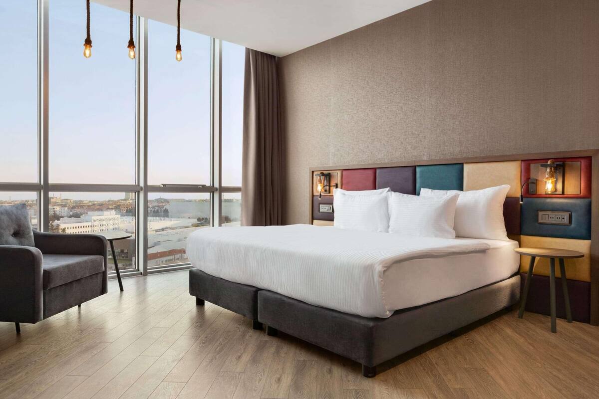 A modern hotel room with a large bed, a small table, and a chair. The room has large windows offering a city view.