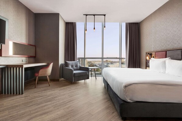 A modern hotel room with a large bed, a desk, a chair, and a view of the city through large windows.