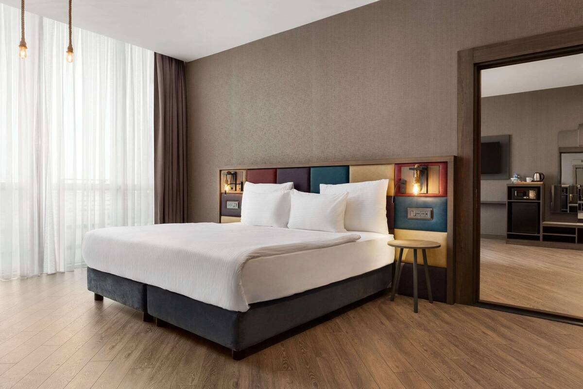 A modern hotel room with a large bed, a bedside table with a lamp, and a view of the kitchen area through an open door.