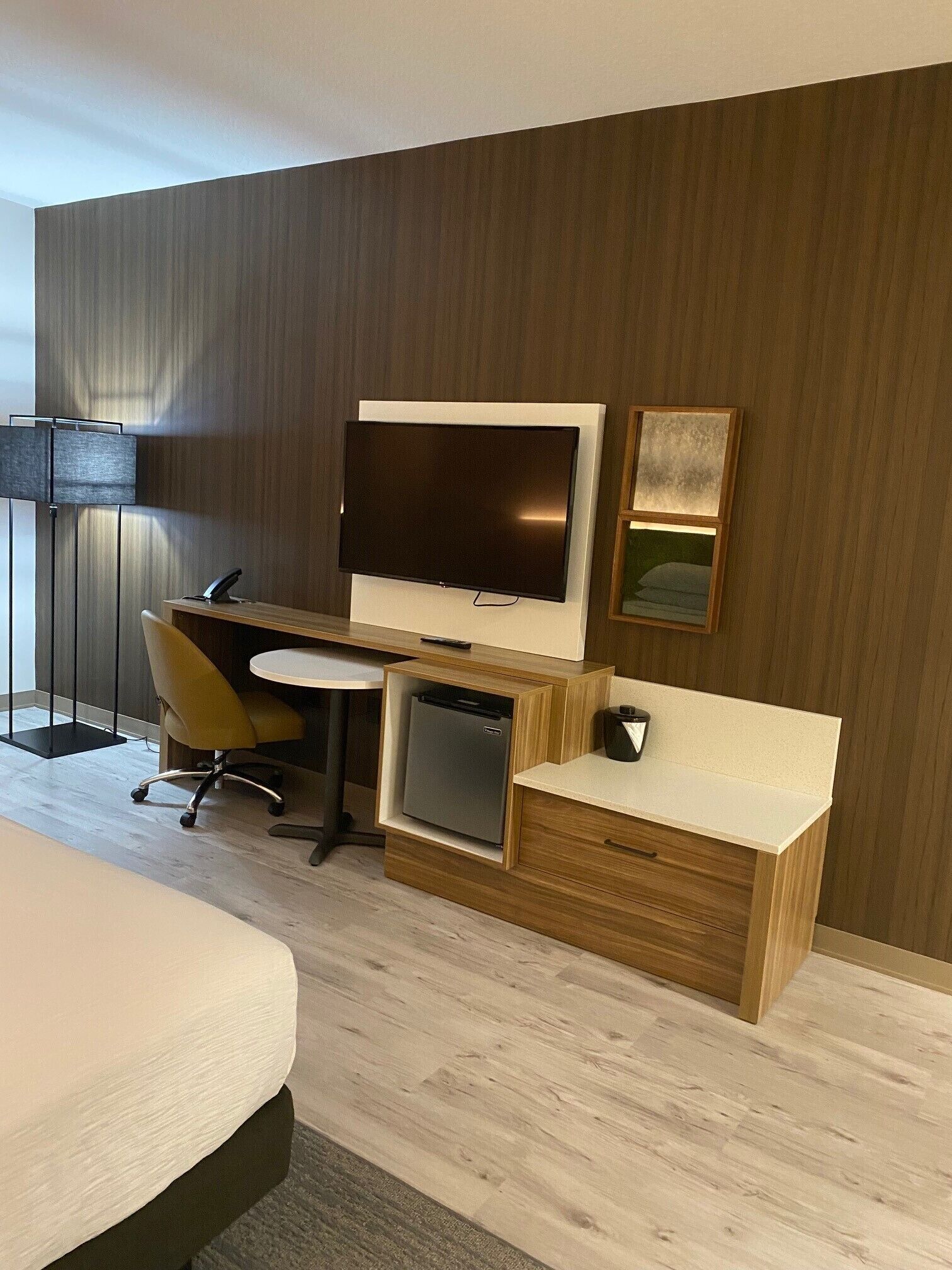 A modern hotel room with a wooden desk, a flat-screen TV, a small refrigerator, a chair, and a bed.