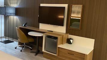 A modern hotel room with a wooden desk, a flat-screen TV, a small refrigerator, a chair, and a bed.