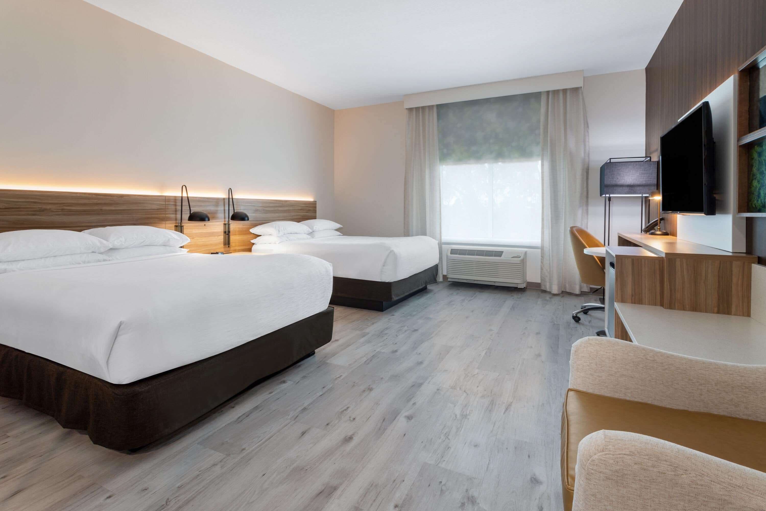 Quarto de hotel com duas camas, uma escrivaninha, uma cadeira, uma televisão e uma janela grande.