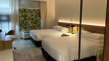 A modern hotel room with two beds, a desk, and a large window with a view of greenery.