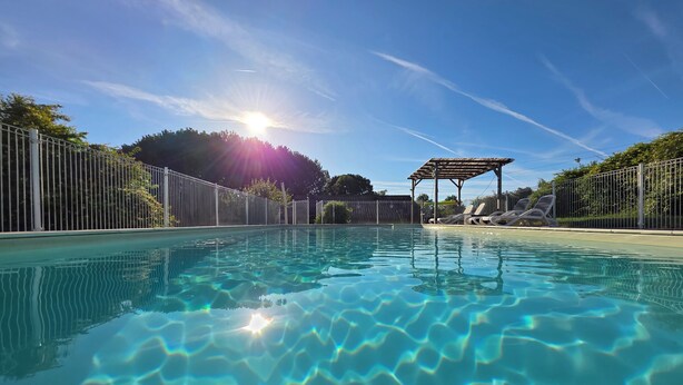 A clear swimming pool with a sunlit sky, a fenced area, and a pergola with lounge chairs.