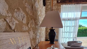 A rustic interior with a stone wall, a wooden table with a woven placemat, a candle, a lamp, and a sign reading 'Gîte du ROC'.