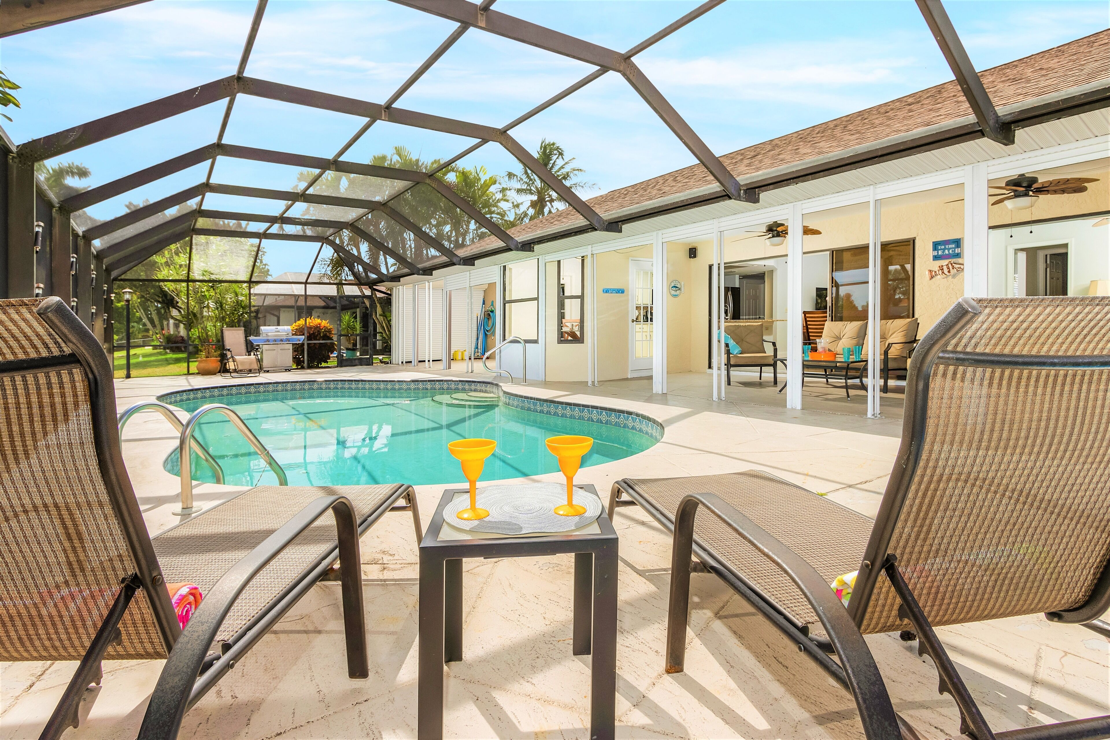 Uma área de piscina descoberta coberta, com espregui çadeiras e uma mesa com duas bebidas amarelas.