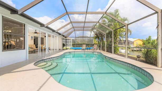 A covered swimming pool with a glass ceiling, surrounded by palm trees and a view of a lake. - Family-friendly villa w/bikes & pool table; Gulf access! Villa Lilly - SWFL Rentals (Cape Coral)
