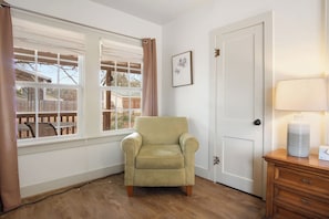 Een kamer met een lichtgekleurde fauteuil, een deur, een raam met uitzicht op een achtertuin en een lamp op een nachtkastje van hout.