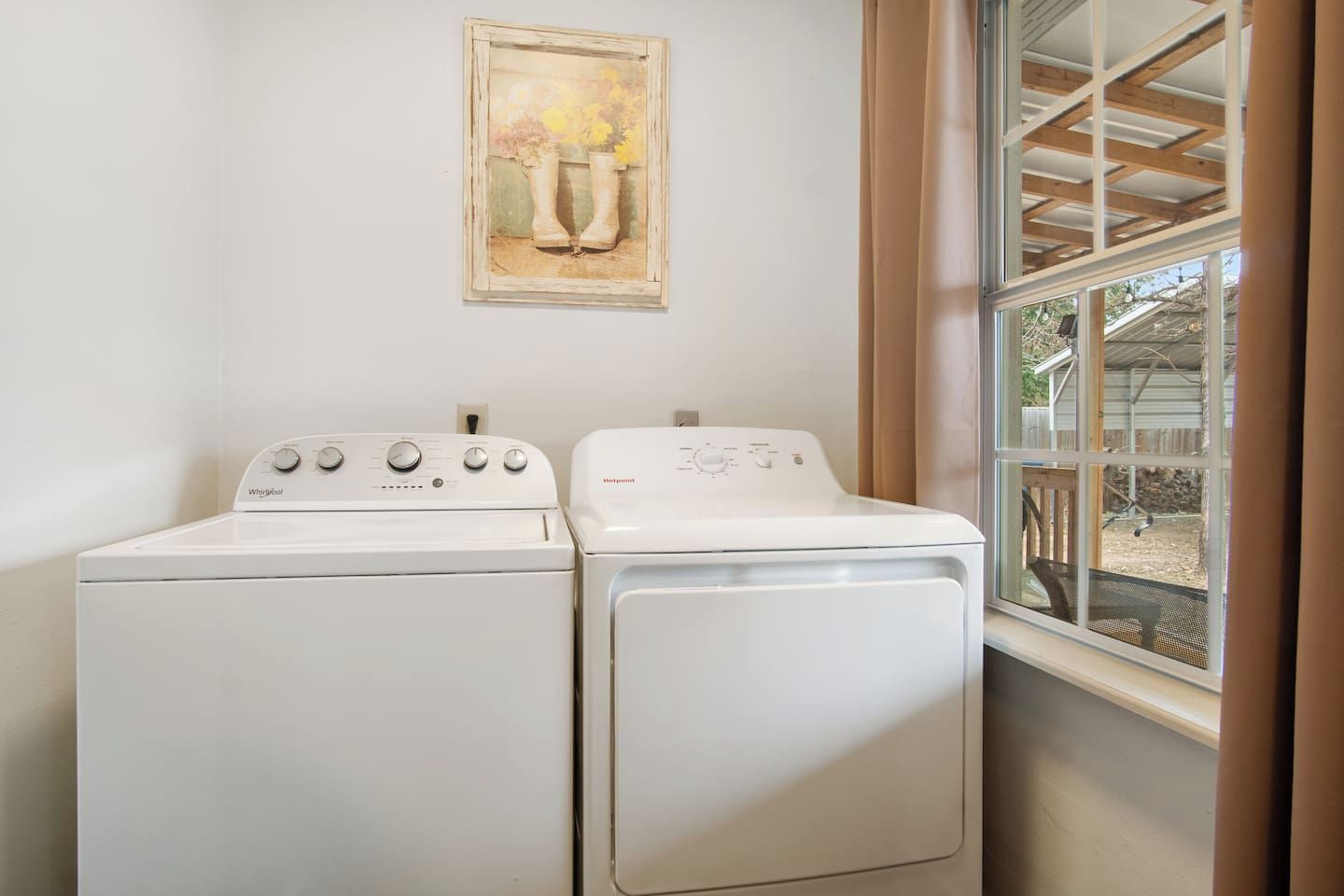 A laundry room with two white Whirlpool washers and dryers, a painting above the dryers, and a window with a view of a wooden deck.
