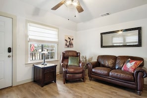A living room with a brown leather sofa, a leather armchair, a wooden side table with a lamp, and a large window with blinds. - Cherry Street | Near Main | Gas BBQ | Couples (Bandera)