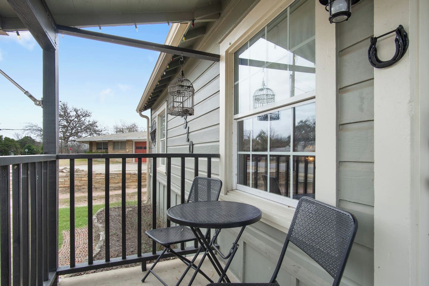 Un balcon avec une table et deux chaises, une fenêtre avec une cage à oiseaux et une vue sur une maison et des arbres.