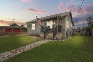 Een huis met twee verdiepingen, een veranda, een vlag en een pad van stenen dat naar de ingang leidt.