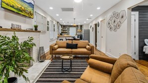 Una sala de estar moderna con un sofá, una mesa de centro y un televisor montado en la pared. Se observa una cocina al fondo, junto a un comedor.