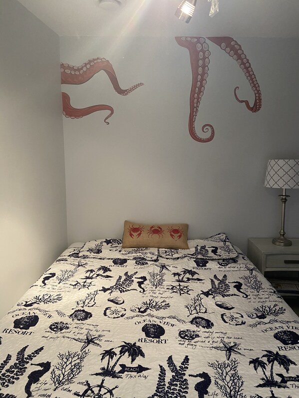 A bedroom with a bed featuring a marine-themed bedspread, a wall with an octopus mural, and a bedside lamp.