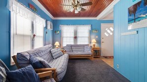 Una stanza con soffitto in legno, due finestre con persiane bianche, un letto con lenzuola a righe, una sedia in legno e un tavolino con una lampada.