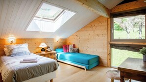 A cozy attic bedroom with a bed, a blue sofa, a small table, and a window overlooking a garden.