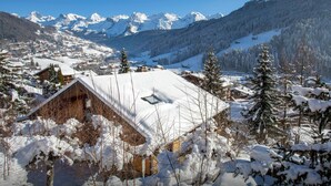 A snow-covered mountain village with traditional wooden houses, a clear blue sky, and a view of snow-capped peaks in the distance.