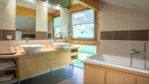 A modern bathroom with a large sink, vanity, and bathtub. It features wooden beams, a large mirror, and a window providing natural light.