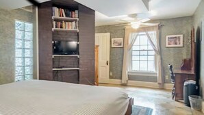 Quarto com uma cama grande, uma estante de livros, um ventilador de teto e uma janela com cortinas.