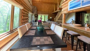 A spacious log cabin interior with a dining area, a stone fireplace, and a large window overlooking a forest.