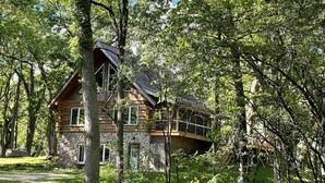 A log cabin with a stone foundation, wooden upper structure, and a balcony, surrounded by dense forest.