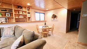 Ein gemütliches Wohnzimmer mit Holzbalkendecke, einem beigefarbenen Sofa, einem Esstisch mit Stühlen und Regalen mit diversen Gegenständen.