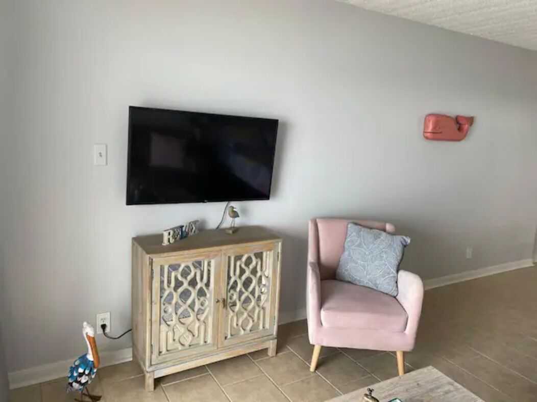 Un salon avec un téléviseur à écran plat fixé au mur, un meuble en bois orné d’objets décoratifs, un fauteuil rose et un petit oiseau décoratif.