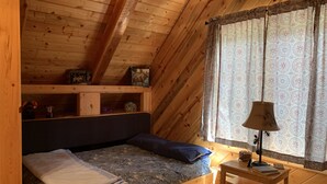 A cozy attic bedroom with a wooden ceiling, a bed with a floral mattress, a small table with a lamp, and a window with patterned curtains.