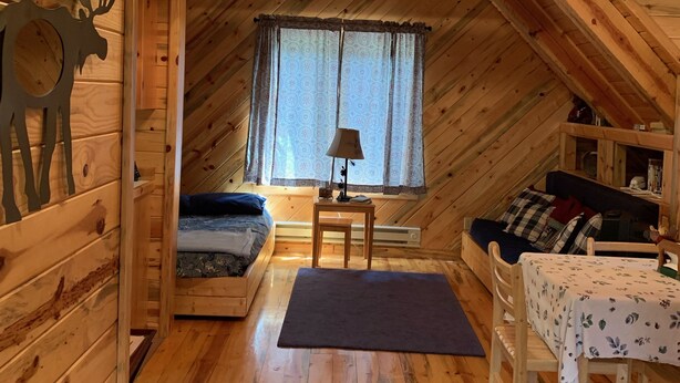 A cozy attic room with wooden walls and a sloped ceiling. It features a bed, a small table, a window with curtains, and a decorative deer head on the wall.