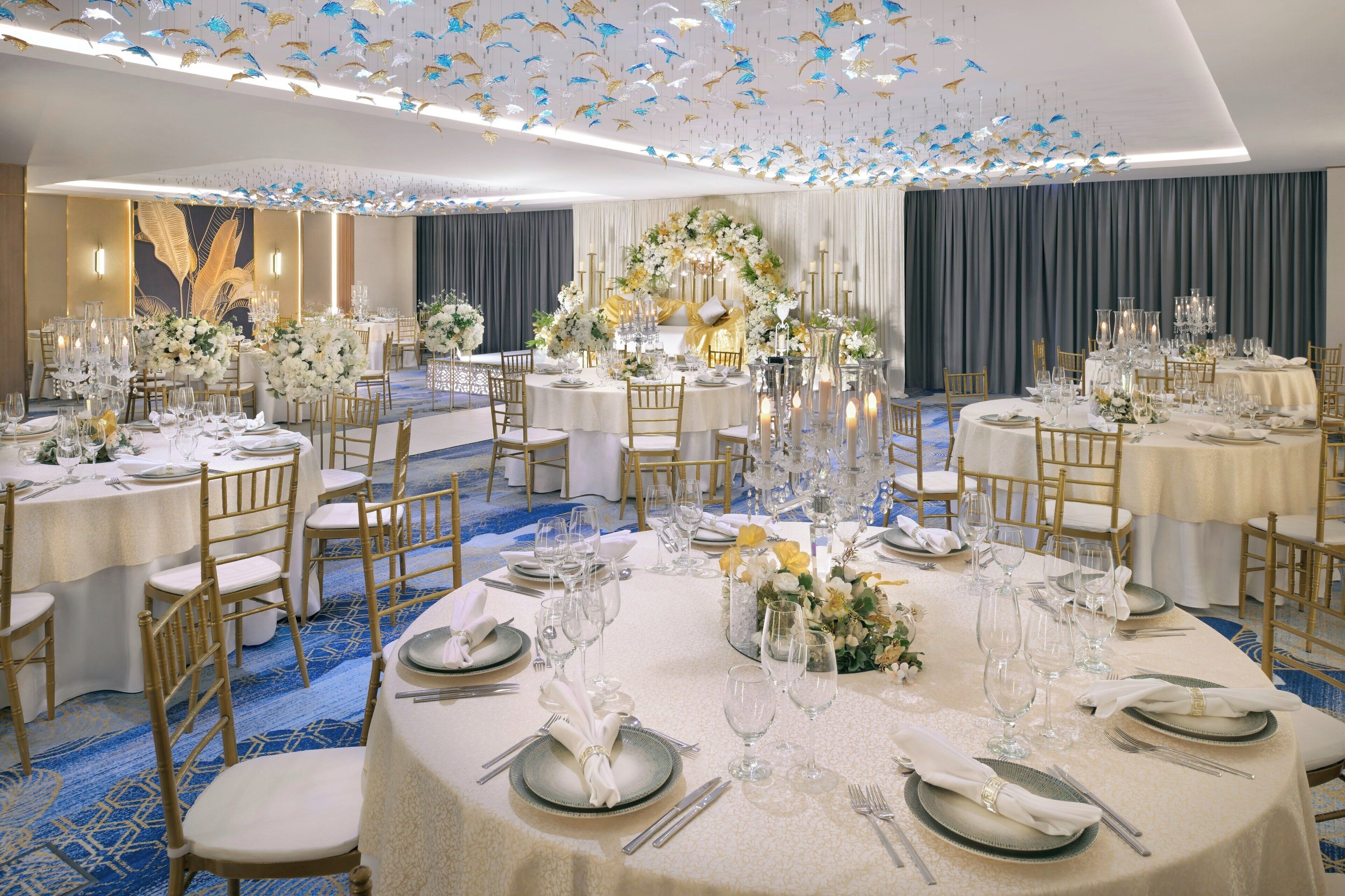 A banquet hall with round tables set for a formal event, featuring gold chairs, white tablecloths, and floral centerpieces.