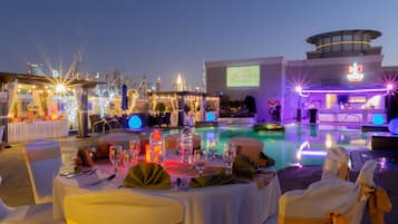 An outdoor pool area with tables set for dining, a pool, and a bar area with a screen displaying a tennis match.