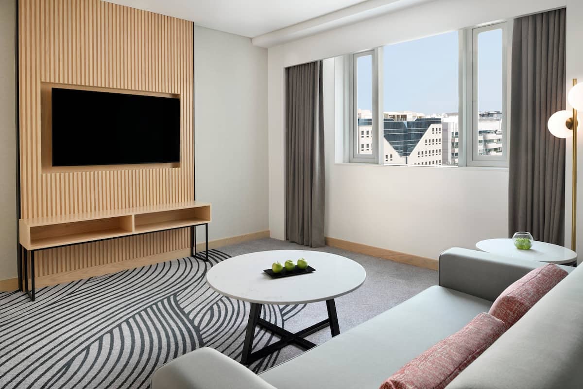 A modern hotel room with a flat-screen TV mounted on a wooden panel, a round white table with a tray of apples, a grey sofa, and a view of city buildings through large windows.