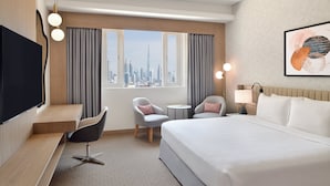 Habitación de hotel con una cama grande, dos sillones, una mesita, un televisor y vistas a la ciudad.