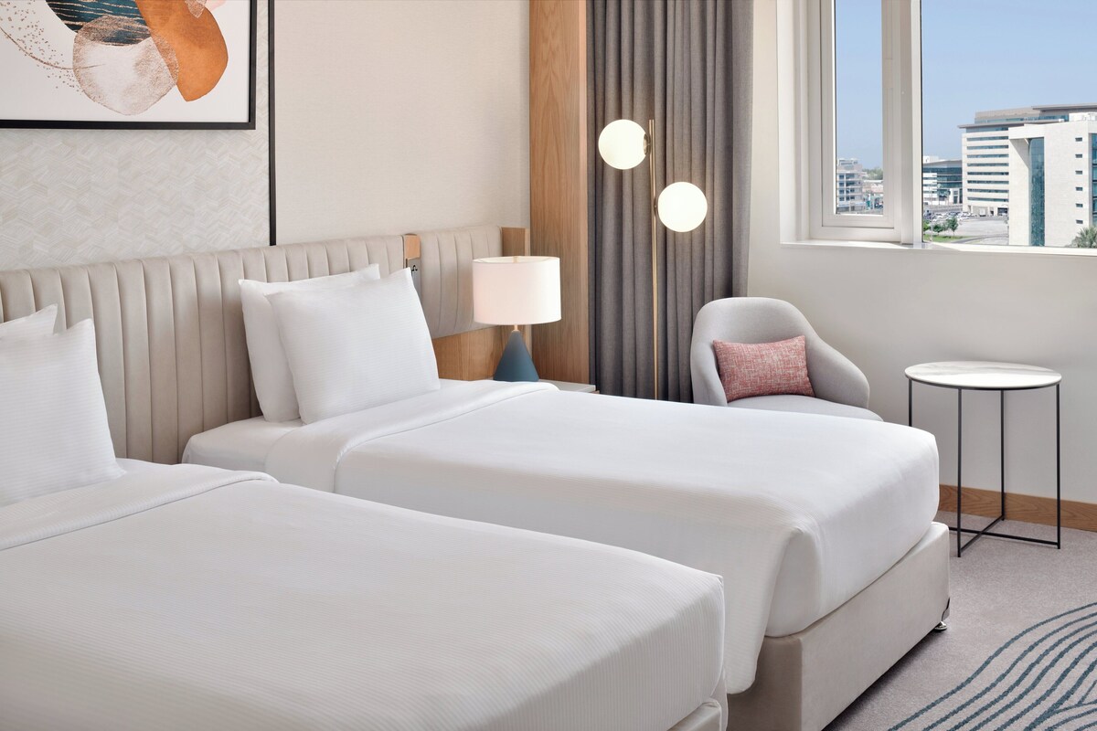 Habitación de hotel con dos camas, una mesita redonda pequeña, una silla, una lámpara y vistas de la ciudad a través de amplios ventanales.