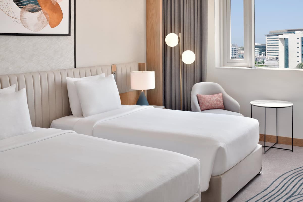 A hotel room with two beds, a small round table, a chair, a lamp, and a view of the city through large windows.
