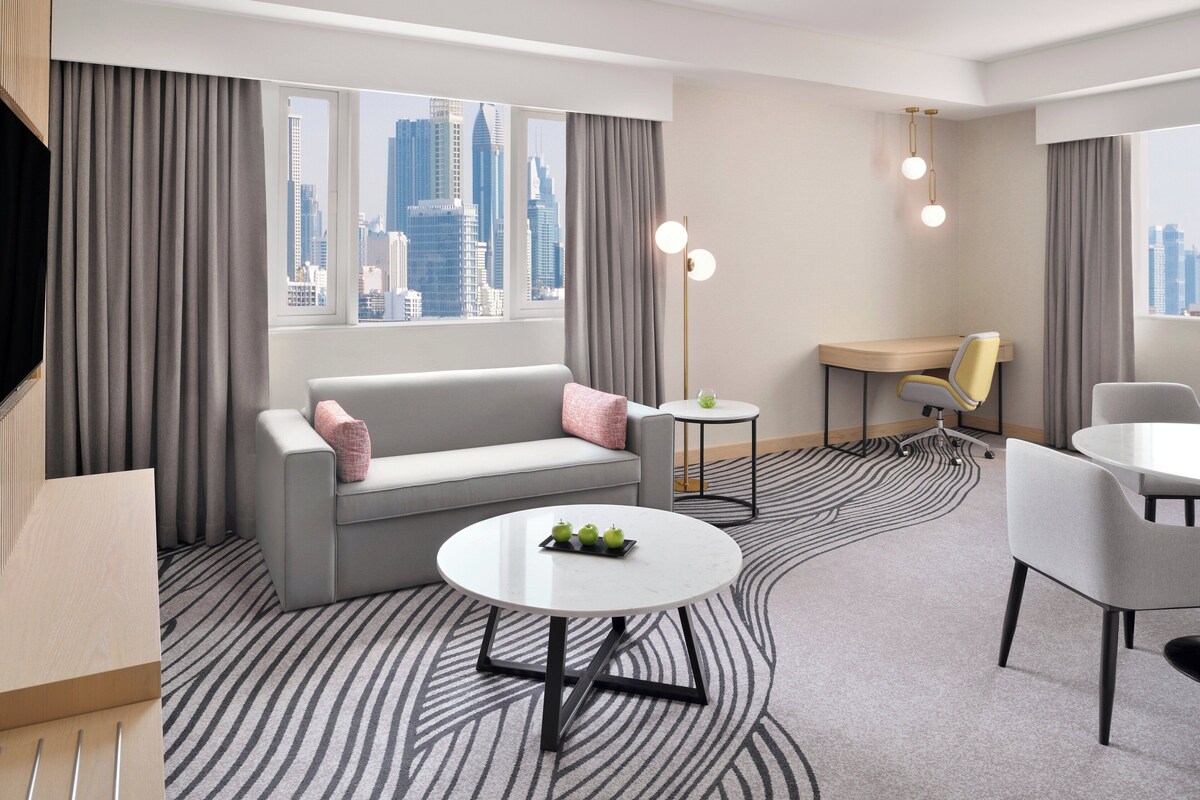 Una habitación de hotel moderna con un sofá gris, una mesa redonda con manzanas, un escritorio y una silla.