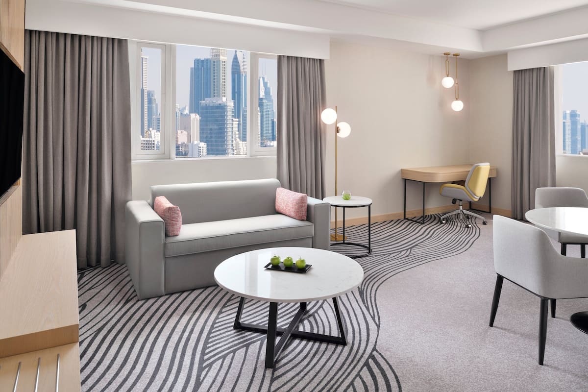 A modern hotel room with a grey sofa, a round table with apples, a desk, and a chair.