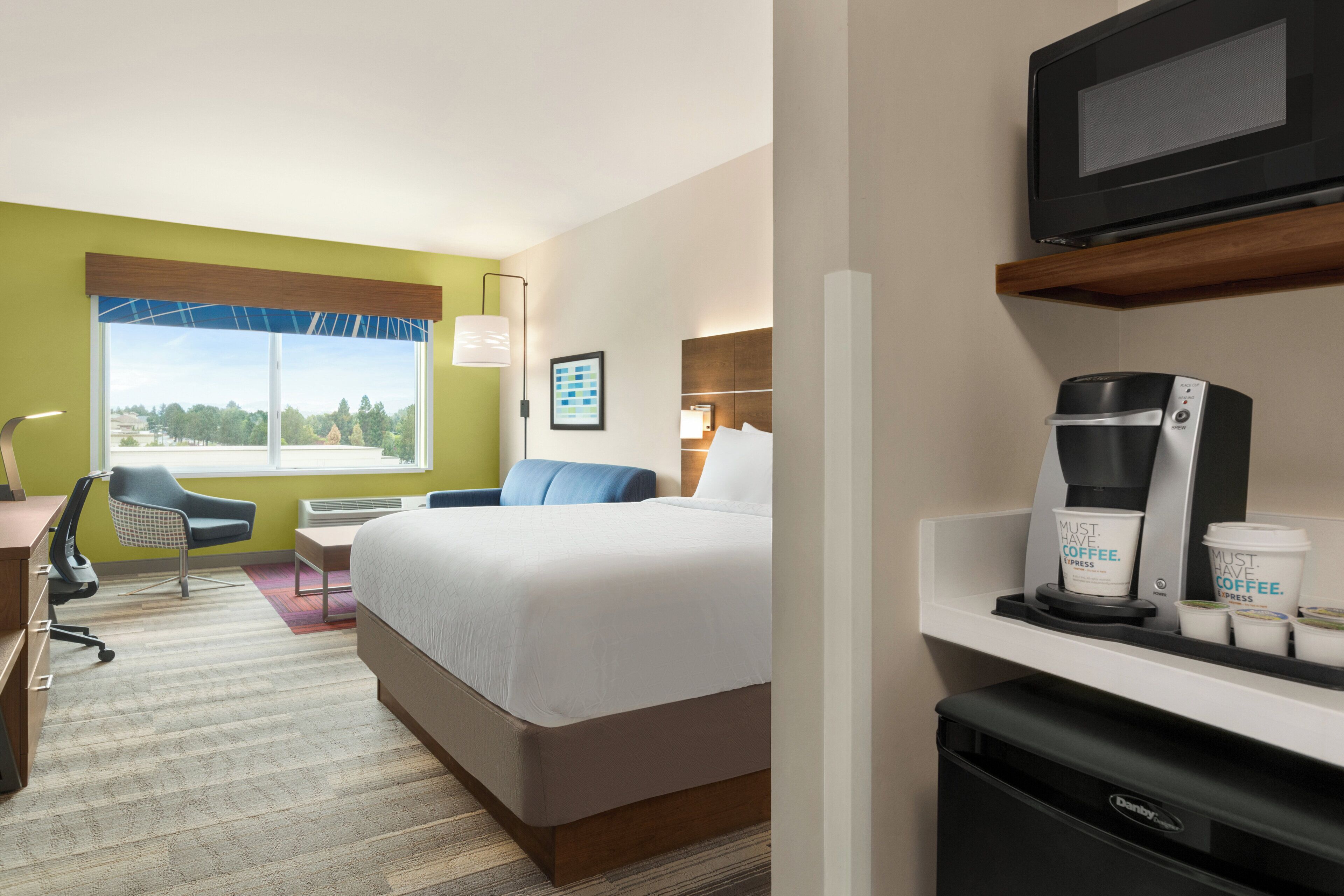 A hotel room with a bed, a desk, a chair, a coffee machine, and a television.