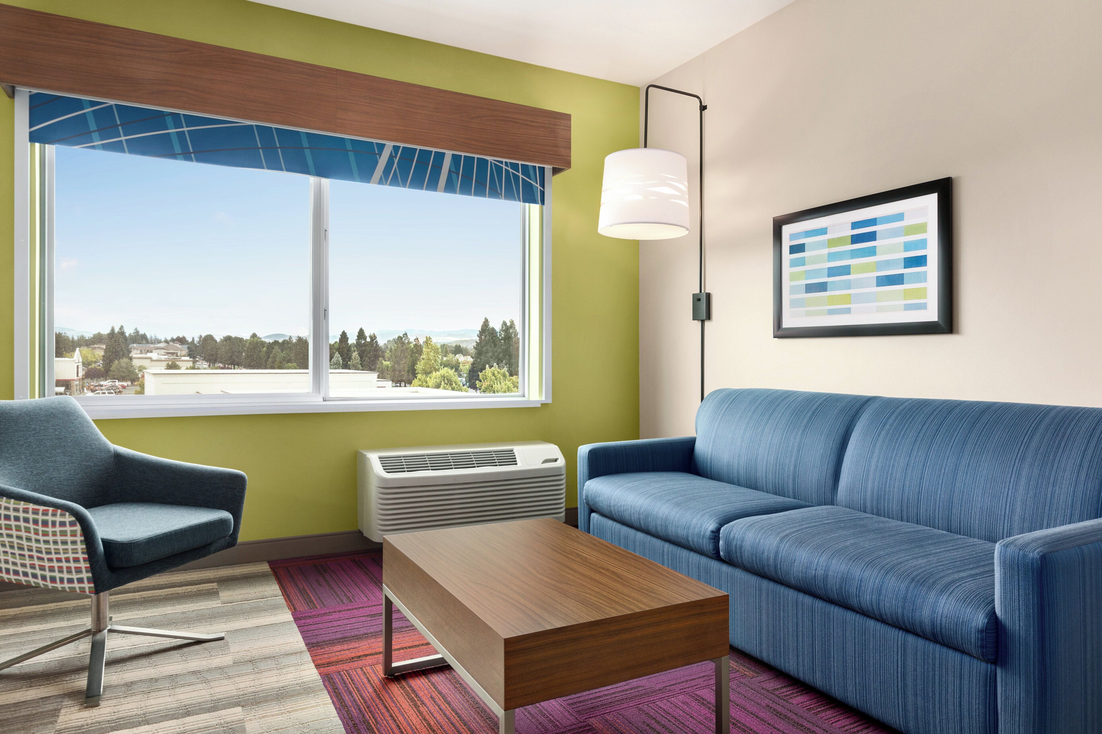 Ein modernes Hotelzimmer mit einem großen Fenster, einem blauen Sofa, einem Holztisch und einem gemusterten Teppich.