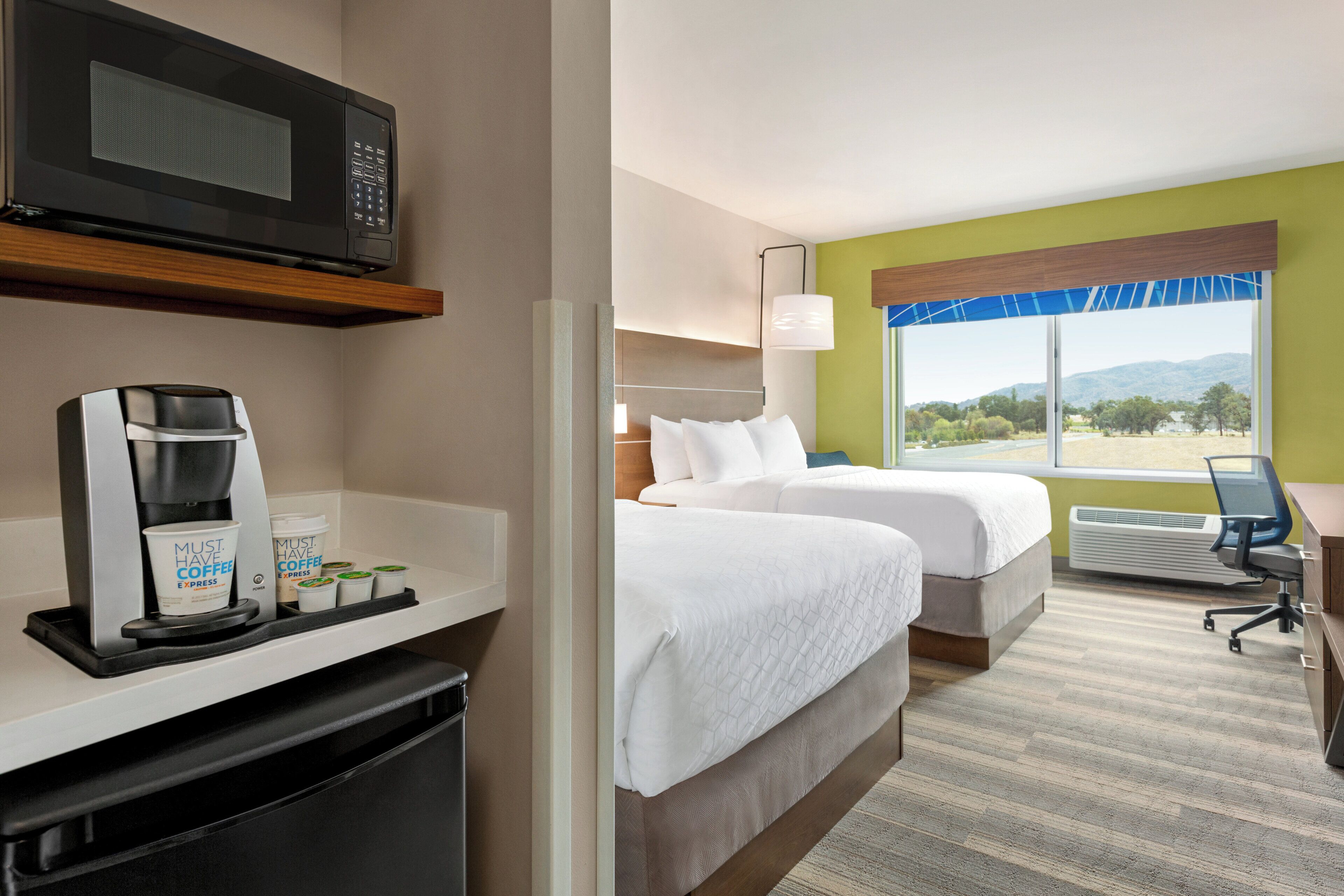 A hotel room with a kitchenette, two beds, a desk, a chair, a window with a view, and a microwave.