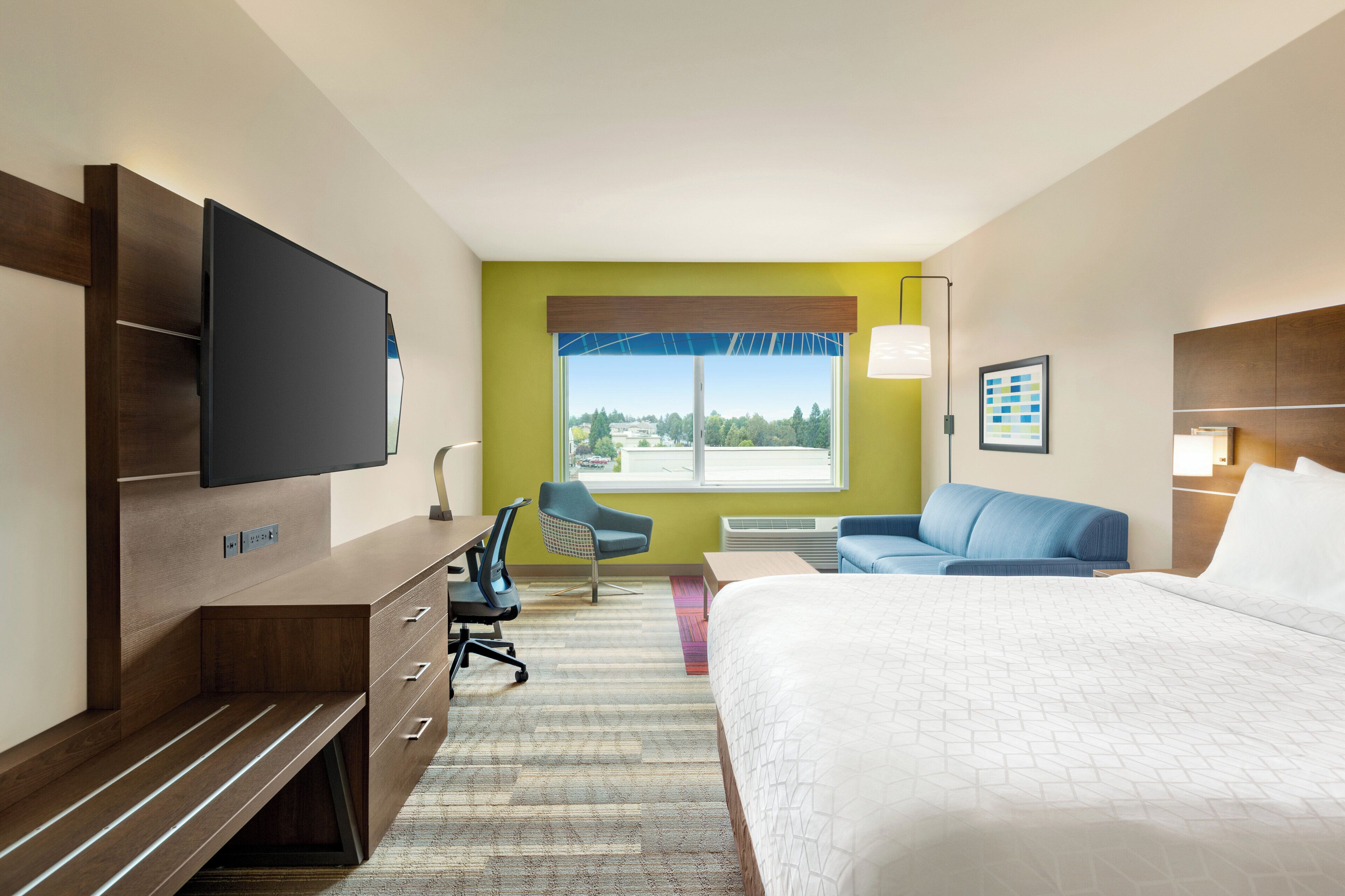 A hotel room with a bed, a desk with a chair, a television, a window with a view, and a sofa.