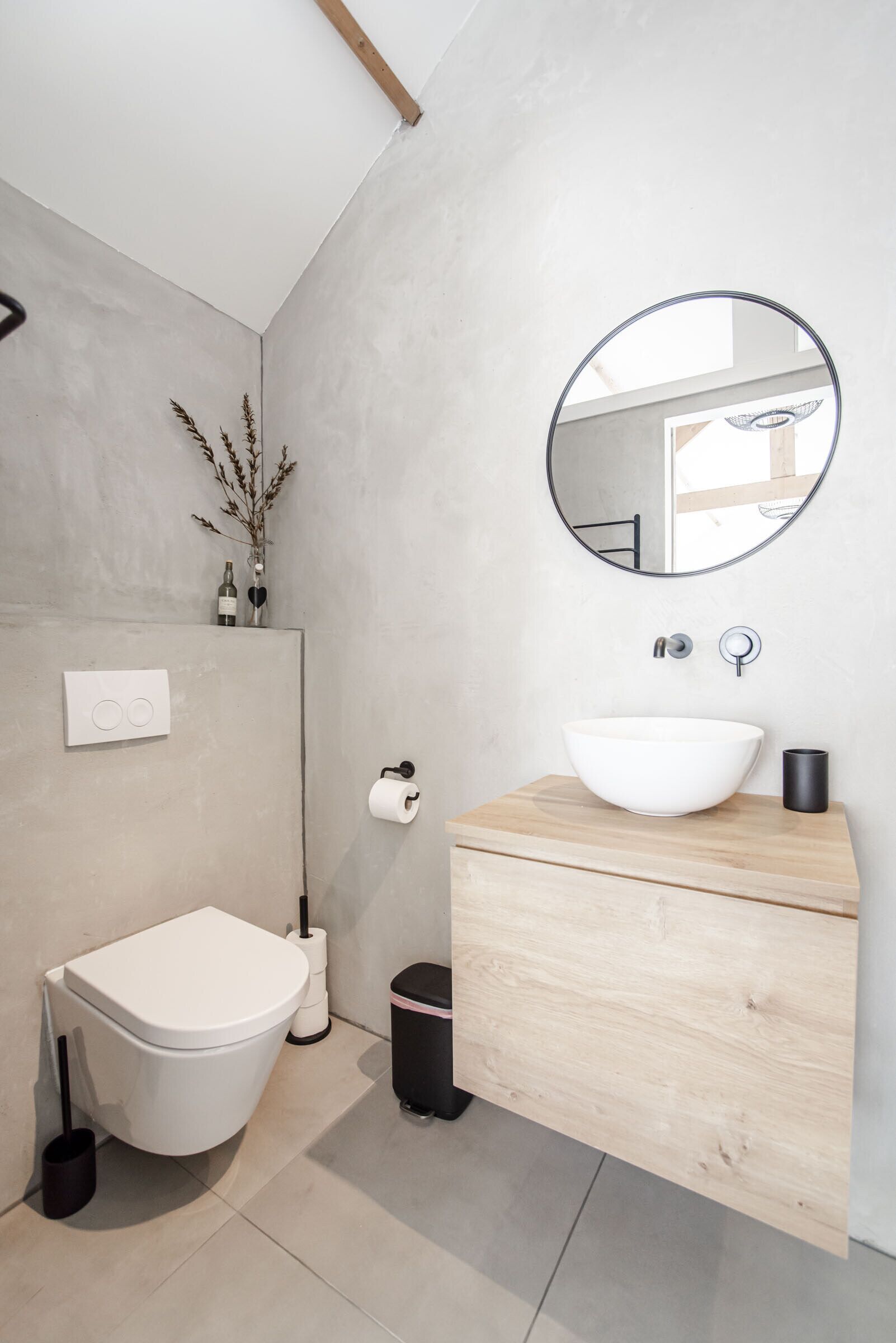 A modern bathroom with a white toilet, a wooden vanity, a round mirror, and a white sink.