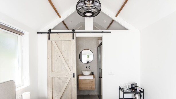 A modern bedroom with a large bed, a wooden door, a small bathroom with a sink, and a black shelf with toiletries. - Be So Suite : Perfect getaway for 2 (Zandvoort)