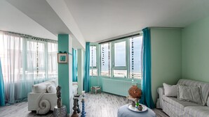 Una habitación con una pared azul claro, un sofá blanco, un reposapiés azul y vistas al mar a través de amplias ventanas.