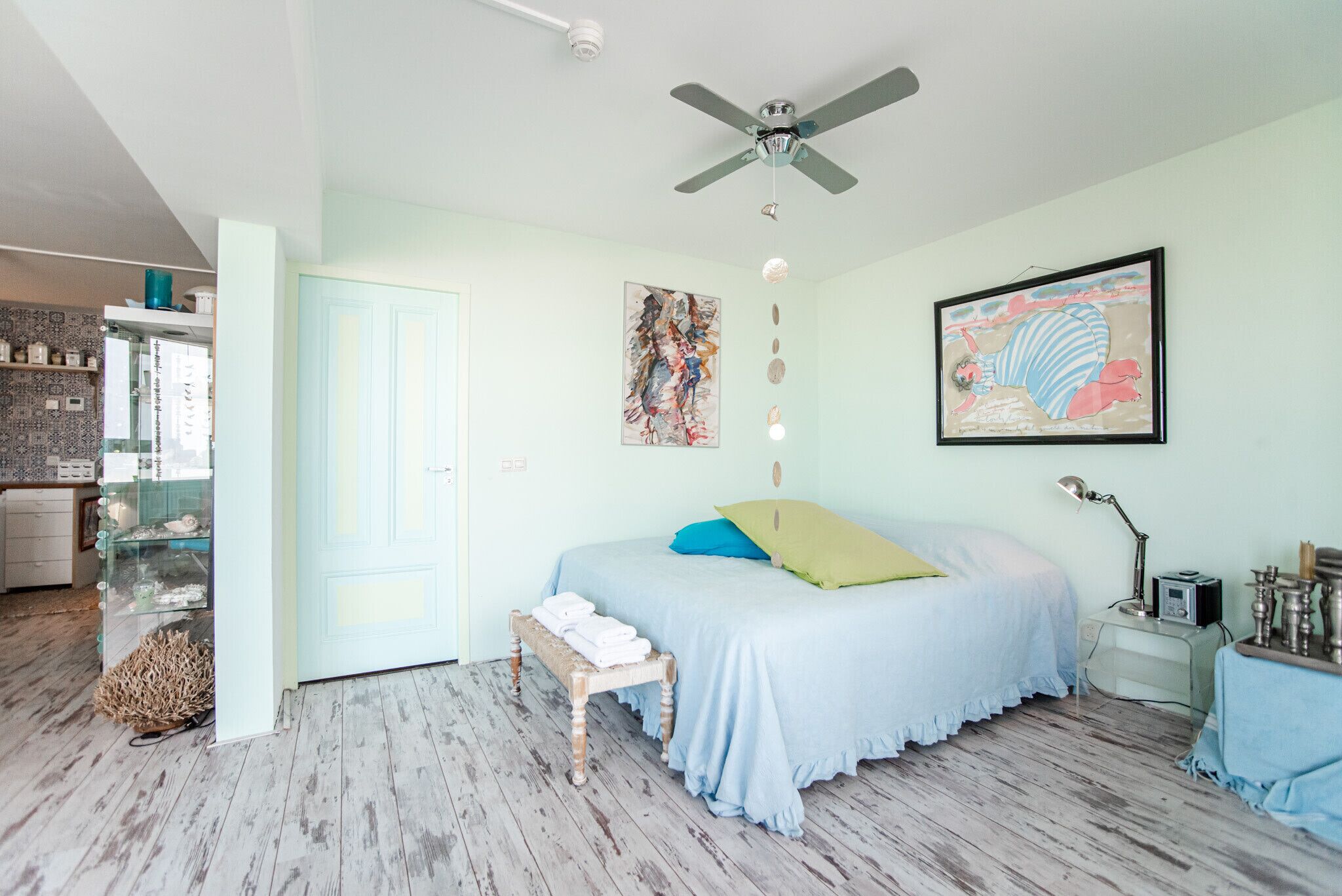 A bedroom with a bed, a ceiling fan, artwork, a bedside table with a lamp, and a window.