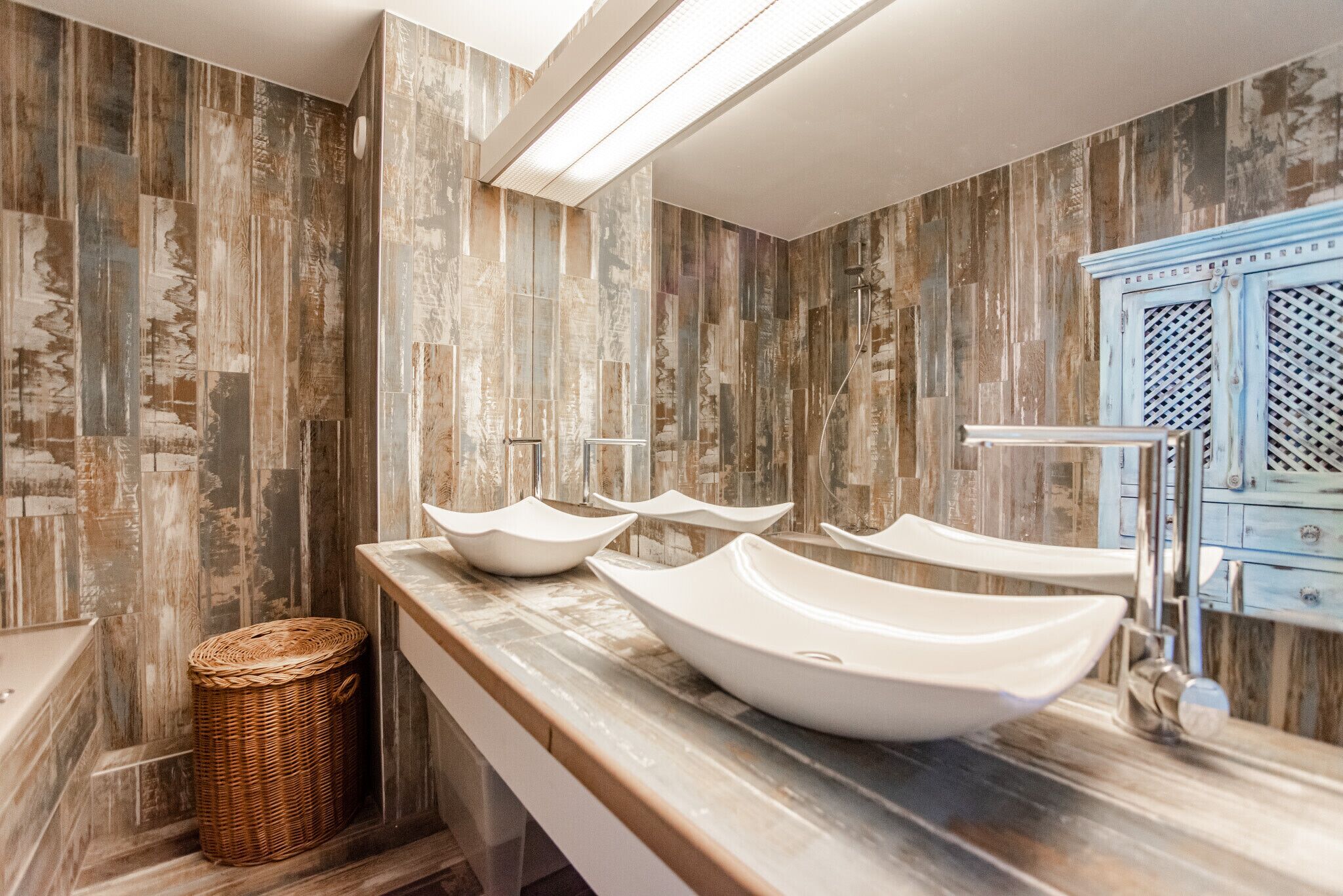 A bathroom with a rustic wood panel wall, a long vanity with three white vessel sinks, and a blue cabinet with a lattice design.