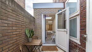Une terrasse extérieure avec un mur en briques, une table en bois, un pot de fleurs et une porte donnant sur un espace intérieur.